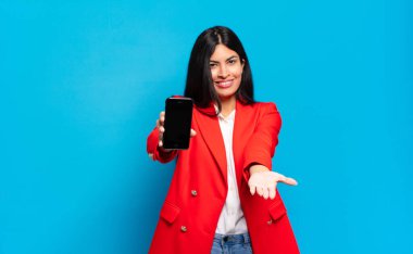 Genç İspanyol kadın mutlu bir şekilde gülümsüyor arkadaş canlısı, kendinden emin, olumlu bakışlı, bir nesne ya da konsept sunuyor ve gösteriyor. telefon ekran kopyalama alanı