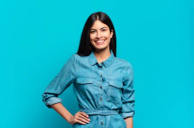 Genç Latin asıllı sıradan bir kadın mutlu bir şekilde gülümsüyor, eli kalçasında ve kendinden emin, olumlu, gururlu ve arkadaş canlısı.