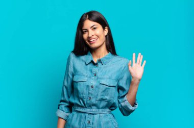 Genç Latin asıllı sıradan bir kadın mutlu ve neşeli bir şekilde gülümsüyor, el sallıyor, sizi selamlıyor ya da veda ediyor.