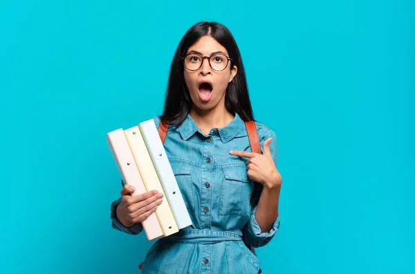 young hispanic student woman looking shocked and surprised with mouth wide open, pointing to self