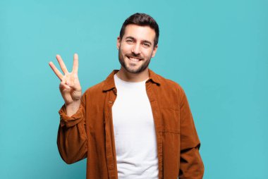 Yakışıklı sarışın adam gülümsüyor ve arkadaş canlısı görünüyor. Üç ya da üç numarayı gösteriyor. Elleri önde, geriye doğru sayıyor.