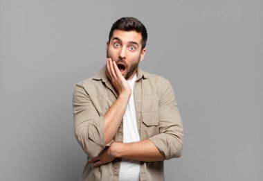 Yakışıklı sarışın adam şok ve inançsızlık içinde ağzı açık, elleri yanağında ve kolu çapraz, sersemlemiş ve şaşırmış hissediyor.