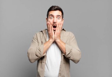 Yakışıklı sarışın adam şok olmuş ve korkmuş hissediyor, ağzı açık ve elleri yanaklarında korkmuş görünüyor.