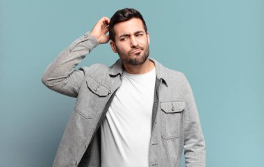 Yakışıklı, sarışın, kafası karışmış, kafası karışmış, başını kaşıyan ve yan tarafa bakan bir adam.