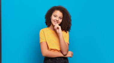  Mutlu görünüyor ve gülümsüyor, elleri çenesinde, merak ediyor ya da bir soru soruyor, seçenekleri karşılaştırıyor.