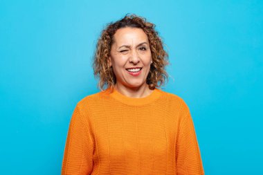 Orta yaşlı bir kadın mutlu ve arkadaş canlısı görünüyor, gülümsüyor ve sana olumlu bir tavırla göz kırpıyor.