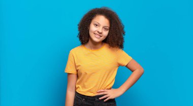  Mutlu bir şekilde gülümsüyor, eli kalçasında ve kendinden emin, olumlu, gururlu ve arkadaş canlısı bir tavırla.