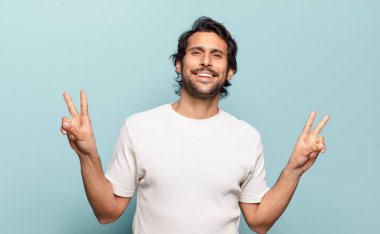 Genç ve yakışıklı Hintli adam gülümsüyor ve mutlu görünüyor, arkadaş canlısı ve tatmin olmuş, iki eliyle zafer veya barış işareti yapıyor.