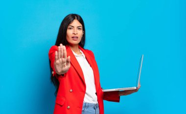 Genç İspanyol kadın ciddi, sert, hoşnutsuz ve kızgın görünüyor. Avuç içinin açık olduğunu göstererek dur hareketi yapıyor. dizüstü bilgisayar