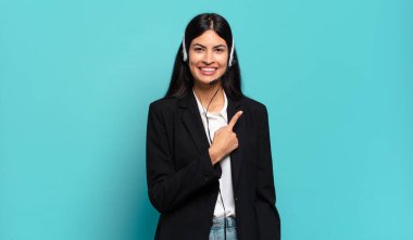 Genç İspanyol pazarlamacı kadın neşeli bir şekilde gülümsüyor, mutlu hissediyor ve yan ve yukarı doğru işaret ediyor, fotokopi uzayındaki nesneleri gösteriyor.