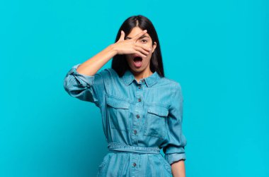 Genç Latin asıllı sıradan bir kadın şok olmuş, korkmuş ya da korkmuş görünüyor. Yüzü elle kaplanmış ve parmaklarının arasından gizlice bakıyor.