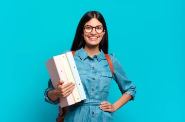 Genç İspanyol öğrenci kadın mutlu bir şekilde gülümsüyor elleri kalçasında ve kendinden emin, olumlu, gururlu ve arkadaş canlısı bir tavırla.