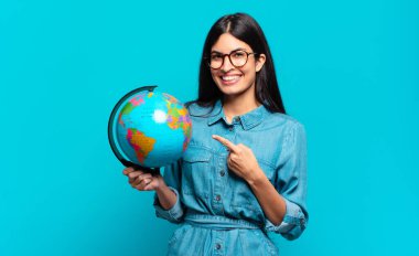 Genç İspanyol kadın neşeli bir şekilde gülümsüyor, mutlu hissediyor ve kenarı ve yukarıyı işaret ediyor, fotokopi uzayında nesneler gösteriyor. Toprak gezegeni konsepti