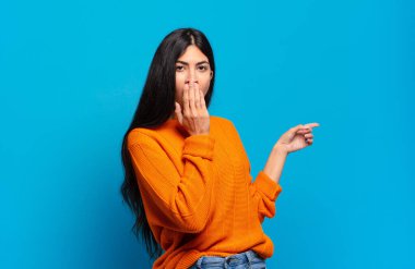 Genç ve güzel İspanyol kadın mutlu, şok olmuş ve şaşırmış hissediyor. Ağzını eliyle kapatıyor ve yanal fotokopi alanını işaret ediyor.