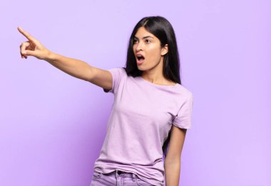 Genç, güzel, İspanyol bir kadın şaşkın ve şaşırmış hissediyor. Şaşkın ve ağzı açık bir şekilde şaşkınlıkla yukarı bakıyor ve işaret ediyor.