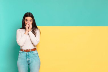 Genç, güzel bir kadın mutlu, neşeli, şanslı ve iki eliyle ağzını kapatırken şaşırmış görünüyor. Konseptinizi yerleştirmek için boşluk kopyalayın