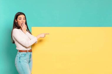 Genç, güzel bir kadın kendini mutlu, şok olmuş ve şaşırmış hissediyor. Ağzını eliyle kapatıyor ve yanal fotokopi alanını işaret ediyor. Konseptinizi yerleştirmek için boşluk kopyalayın