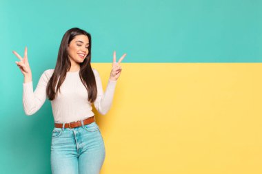Genç, güzel bir kadın gülümsüyor ve mutlu görünüyor. Dost canlısı ve memnun. İki elinle zafer ya da barış işareti yapıyor. Konseptinizi yerleştirmek için boşluk kopyalayın