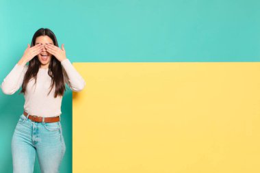 Genç ve güzel bir kadın gülümsüyor ve mutlu hissediyor. İki eliyle gözlerini kapatıyor ve inanılmaz bir sürpriz bekliyor. Konseptinizi yerleştirmek için boşluk kopyalayın