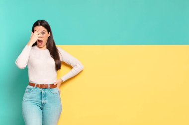 young pretty woman looking shocked, scared or terrified, covering face with hand and peeking between fingers. copy space to place your concept