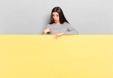 young pretty woman feeling confused, puzzled and insecure, pointing to self wondering and asking who, me?. copy space to place your concept