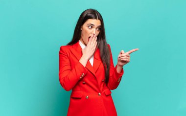 young pretty woman feeling happy, shocked and surprised, covering mouth with hand and pointing to lateral copy space. business concept