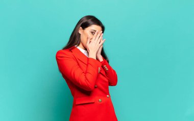 young pretty woman covering face with hands, peeking between fingers with surprised expression and looking to the side. business concept