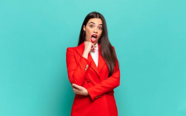 young pretty woman open-mouthed in shock and disbelief, with hand on cheek and arm crossed, feeling stupefied and amazed. business concept