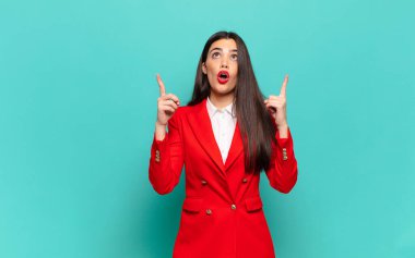 young pretty woman looking shocked, amazed and open mouthed, pointing upwards with both hands to copy space. business concept