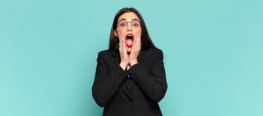 young pretty woman feeling shocked and scared, looking terrified with open mouth and hands on cheeks. business concept