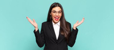 young pretty woman looking happy and excited, shocked with an unexpected surprise with both hands open next to face. business concept