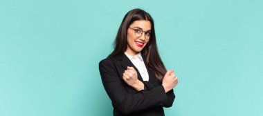 Genç ve güzel bir kadın, neşeli bir şekilde gülümsüyor ve kutluyor, yumruklarını sıkarak ve kollarını kavuşturarak, mutlu ve pozitif hissediyor. iş kavramı