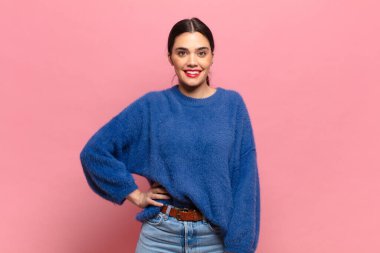 Genç ve güzel bir kadın mutlu bir şekilde gülümsüyor, eli kalçasında ve kendinden emin, pozitif, gururlu ve arkadaş canlısı.