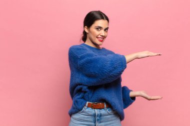 Genç, güzel bir kadın gülümsüyor, mutlu, pozitif ve tatmin olmuş hissediyor, fotokopi alanında nesne veya konsept tutuyor veya gösteriyor