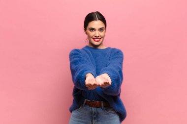 Genç, güzel bir kadın mutlu bir şekilde gülümsüyor. Dost canlısı, kendinden emin, olumlu bakışlı, bir nesne ya da konsept sunuyor ve gösteriyor.