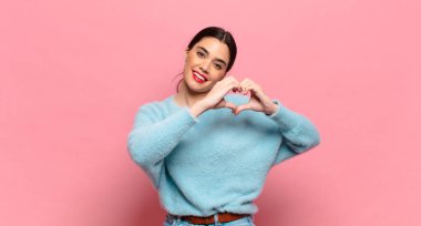 Genç ve güzel bir kadın gülümsüyor ve mutlu, sevimli, romantik ve aşık, iki eliyle kalbi şekillendiriyor