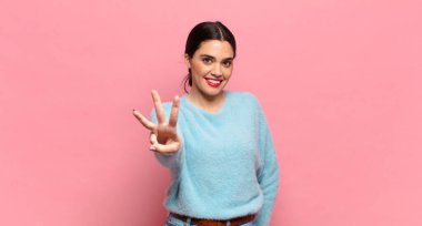 Genç, güzel bir kadın gülümsüyor ve arkadaş canlısı görünüyor. Üç ya da üç numarayı gösteriyor. Elleri önde, geriye doğru sayıyor.