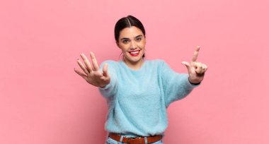 Genç ve güzel bir bayan gülümsüyor ve dost canlısı görünüyor, 7 ya da 7 numarayı gösteriyor, eli önde, geriye doğru sayıyor.