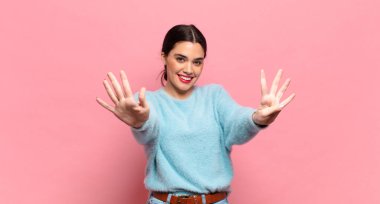 Genç, güzel bir kadın gülümsüyor ve dost canlısı görünüyor. 9 ya da 9 numarayı gösteriyor.