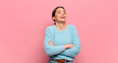 Genç ve güzel bir kadın kollarını kavuşturup mutlu bir şekilde gülüyor, rahat, pozitif ve tatmin edici bir pozla.