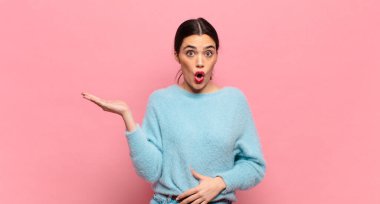 Genç ve güzel bir kadın şaşırmış ve şok olmuş görünüyor, çenesi düşmüş, bir objeyi elinde açık bir elle tutuyor.