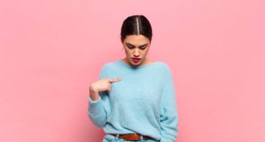 Genç ve güzel bir kadın neşeyle gülümsüyor, yere bakıyor ve göğsüne işaret ediyor.