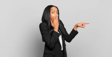young black woman feeling happy, shocked and surprised, covering mouth with hand and pointing to lateral copy space. business concept