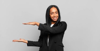 young black woman smiling, feeling happy, positive and satisfied, holding or showing object or concept on copy space. business concept