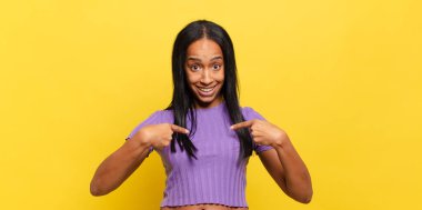 Genç siyahi kadın mutlu, şaşkın ve gururlu hissediyor. Heyecanlı ve şaşkın bir bakışla kendini işaret ediyor.