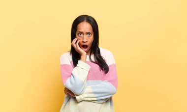 Genç siyahi kadın şok ve inançsızlık içinde, elleri yanak ve kolları çaprazlanmış, sersemlemiş ve şaşırmış hissediyor.