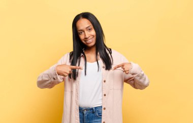 Genç siyah kadın gururlu, pozitif ve rahat görünüyor. İki eliyle göğsünü gösteriyor.