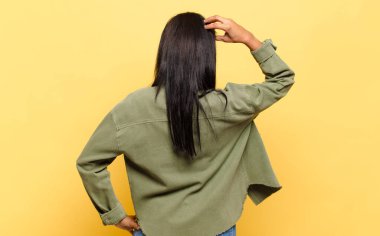 young black woman feeling clueless and confused, thinking a solution, with hand on hip and other on head, rear view