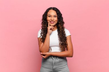 Genç İspanyol kadın mutlu görünüyor ve gülümsüyor, elleri çenesinde, merak ediyor ya da soru soruyor, seçenekleri karşılaştırıyor.