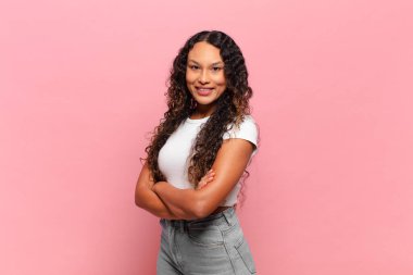 Çapraz kollarıyla kameraya gülümseyen genç İspanyol kadın ve mutlu, kendinden emin, tatmin olmuş bir ifade.